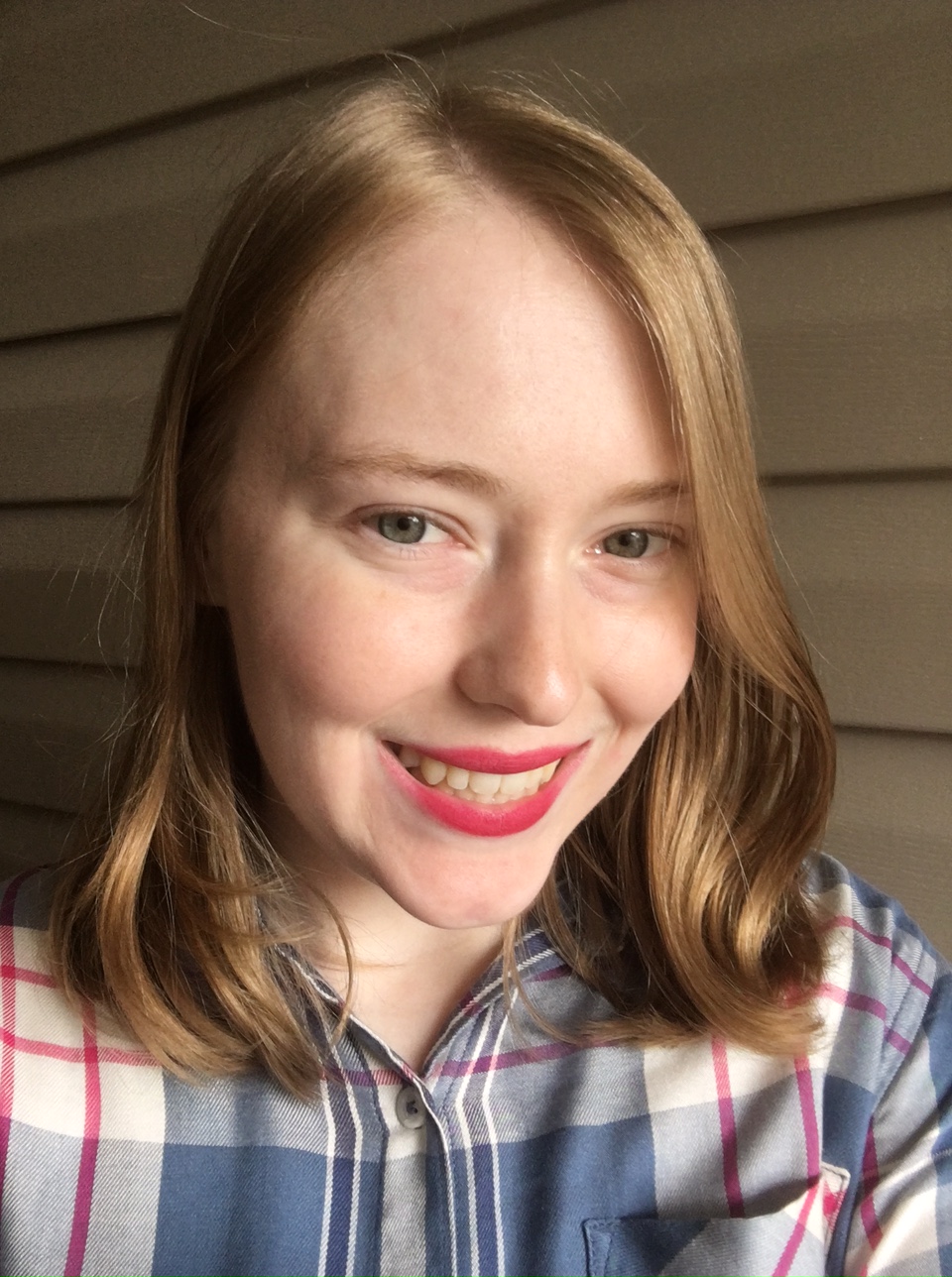 a-woman-with-medium-blonde-hair-and-green-eyes-wearing-a-blue-and-pink-plaid-shirt-and-smiling-at-the-camera