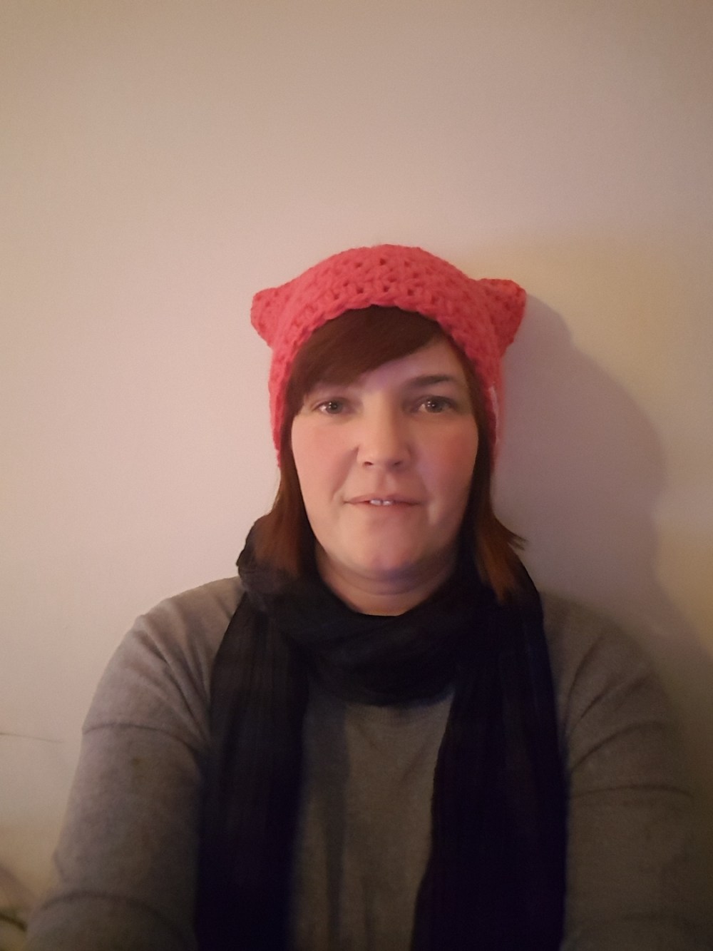 Woman with reddish brown shoulder length hair wearing a bright pink 'pussyhat', grey jumper and dark scarf against an off white wall.