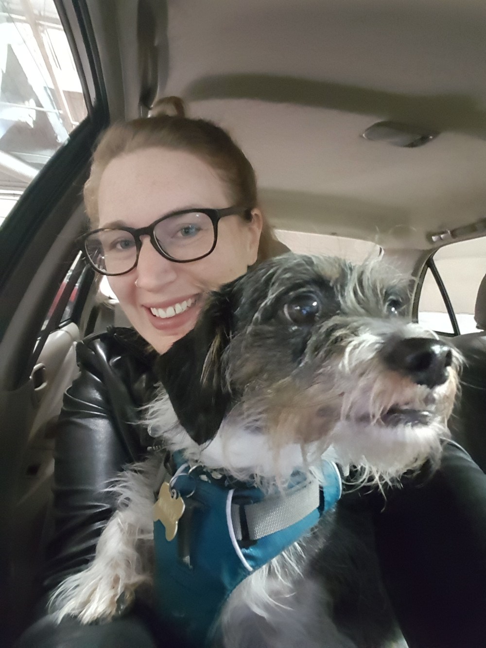 Blonde woman with big glasses, a leather jacket, and an adorable scruffy dog sitting in a car. 