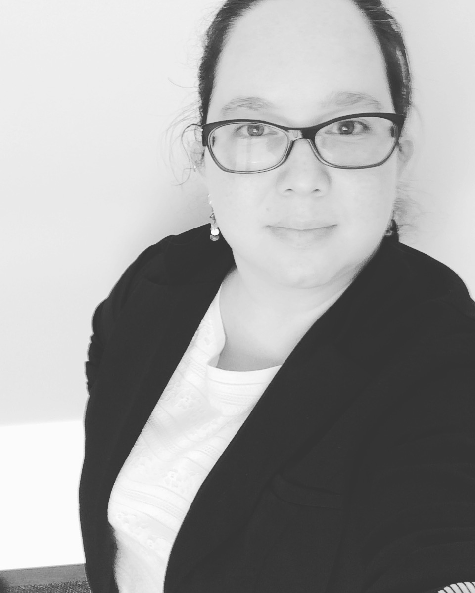 a black and white photo of the head and shoulders of a woman with glasses in a white shirt and black jacket, hair pulled back, looking at the camera.