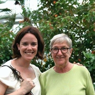 A woman on the left with chin length dark hair in a cream shirt with her arm around a woman with short grey hair wearing glasses and a green shirt.