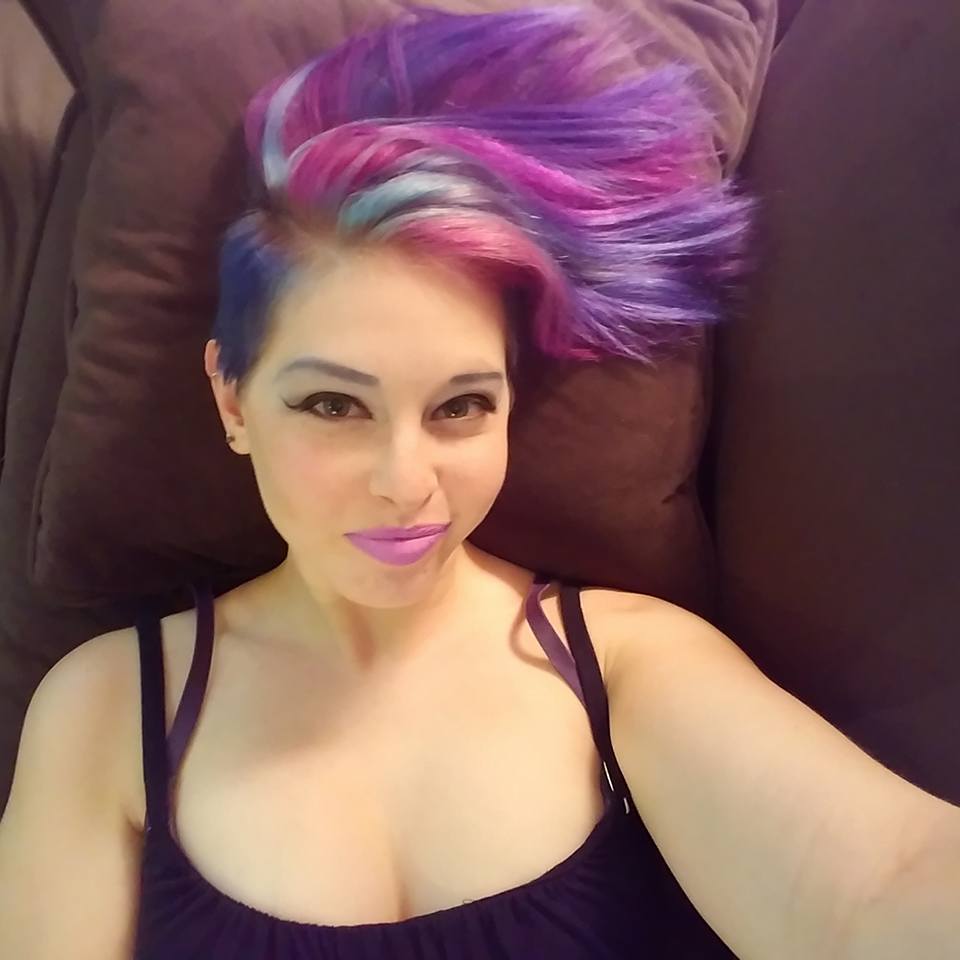 A woman with galaxy hair (blue, purple, turquoise, and pink) lying down on the couch, looking up at the camera with a smile.