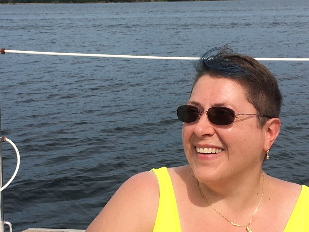 Woman with very short hair in a bright yellow tank top and sunglasses sitting with a lake behind her (and a white cord just behind her head).