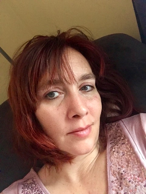 Headshot of a woman with short red hair, looking at the camera and wearing a pink top with sequins.