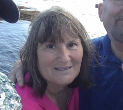 Headshot of Cheryl, who has shoulder-length brown hair. Someone's arm is around her shoulder and she's wearing a bright pink shirt. She's smiling at the camera.