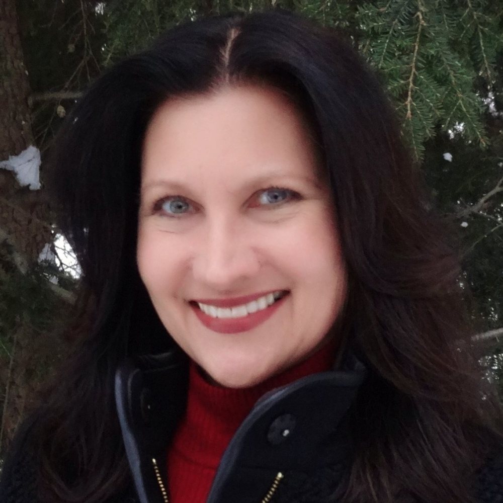 Headshot of Christina outside, in front of an evergreen tree. She has a big smile and is looking directly at the camera.