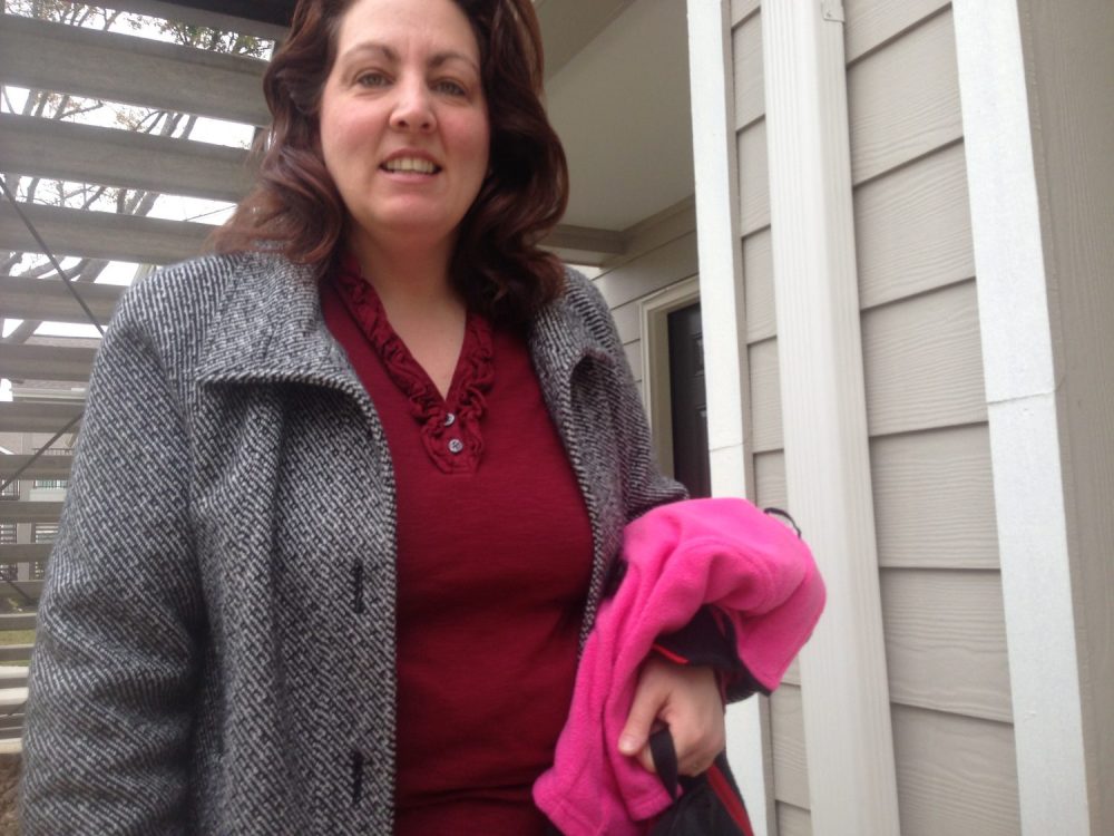 Woman on a porch with a grey coat on, unbuttoned. She wears a red shirt and holds a pink fleece. She's looking and smiling at the camera.