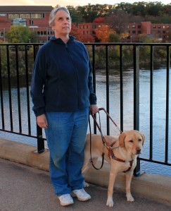 Kathie and her Seeing Eye dog walking across a bridge with fall foliage in the background.