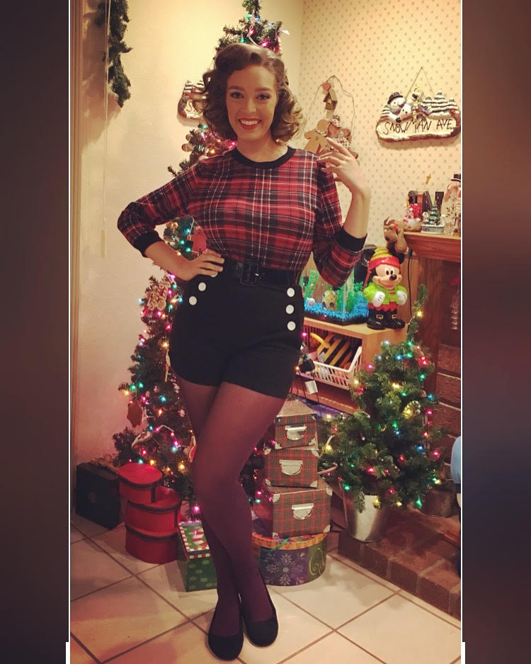 A girl standing in front of a christmas tree with black shorts and a plaid shirt.