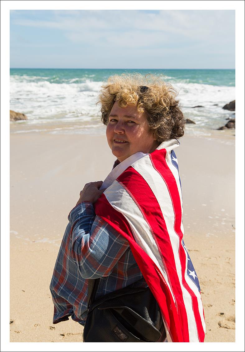 Photo: curly-haired woman by the sea, with an American flag draped over her shoulders.