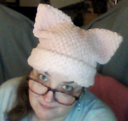 Photo taken from above, a woman smiles up at the camera. She's wearing glasses and a light pink "pussy" hat.