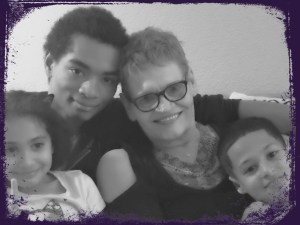 Four people sitting together on a couch, from left to right a girl, a young man, an older woman with short hair and glasses, and a young boy, all embracing