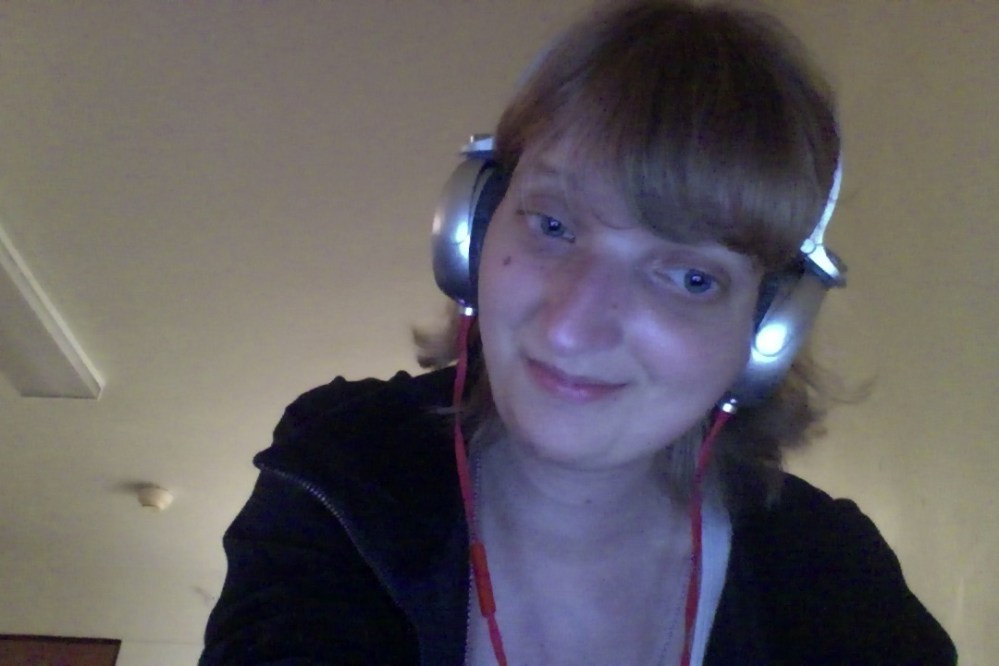 White woman with short blonde hair wearing headphones and a black jacket, smiling and looking slightly away from the camera and to the right. 