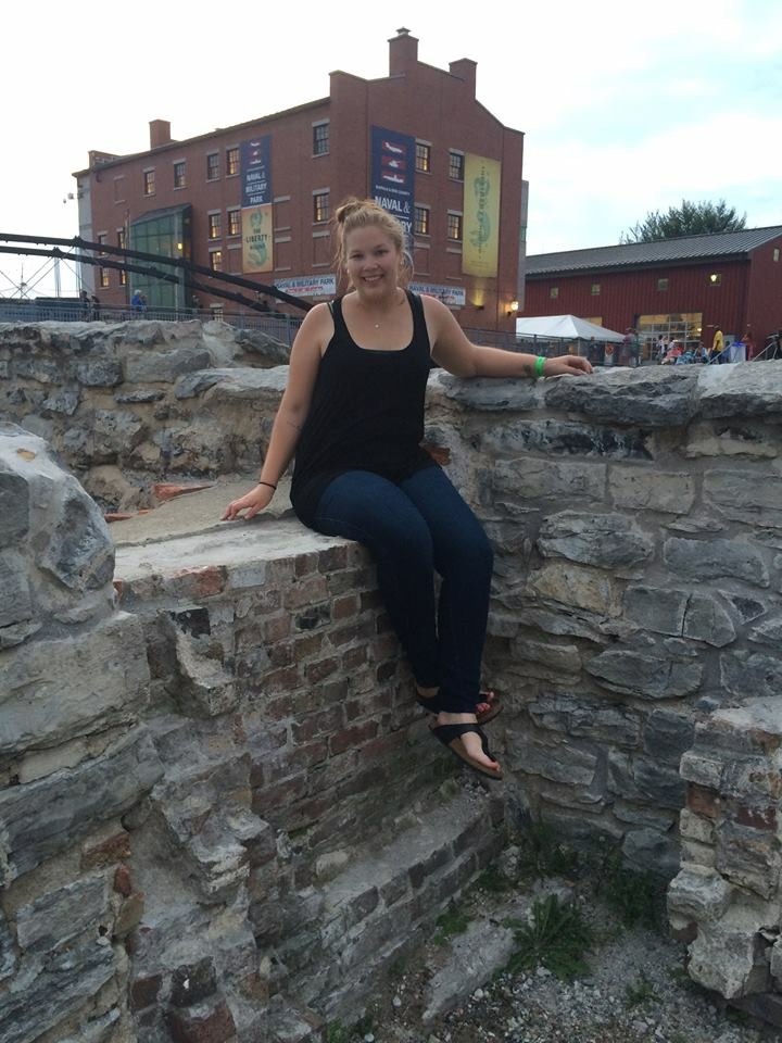 Girl in black tank top and jeans sitting on rock wall in front of brick building