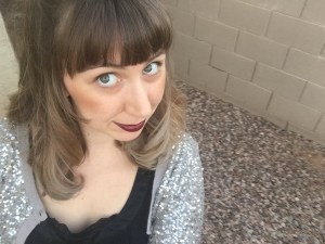 A woman with dark blonde hair and bangs, looking up toward the camera, wearing a gray cardigan and black t-shirt, with cinderblock wall and floor in background