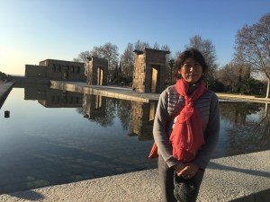 A woman with a red scarf around her neck stands in front of a still reflective pool of water with stone structures behind her.