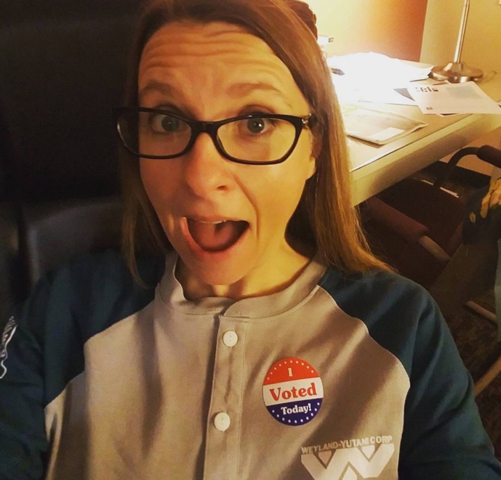 Woman with long, light brown hair wears glasses and looks at the camera with her mouth open. She wears a button-up raglan sleeve shirt with a sticker that says "I Voted."