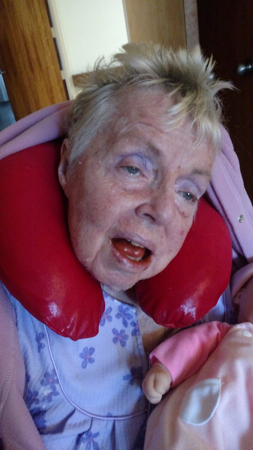 Headshot of an older woman with short blonde hair resting her face on a red neck pillow.