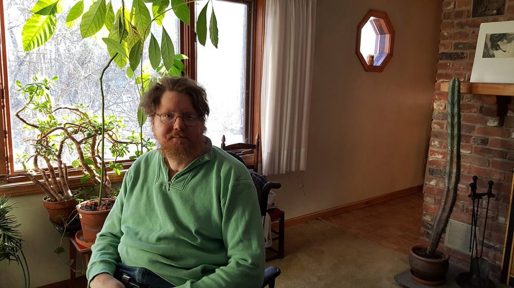 Man with a beard and mustache sitting in front of a window, indoors, with succulent and leafy plants in the background.