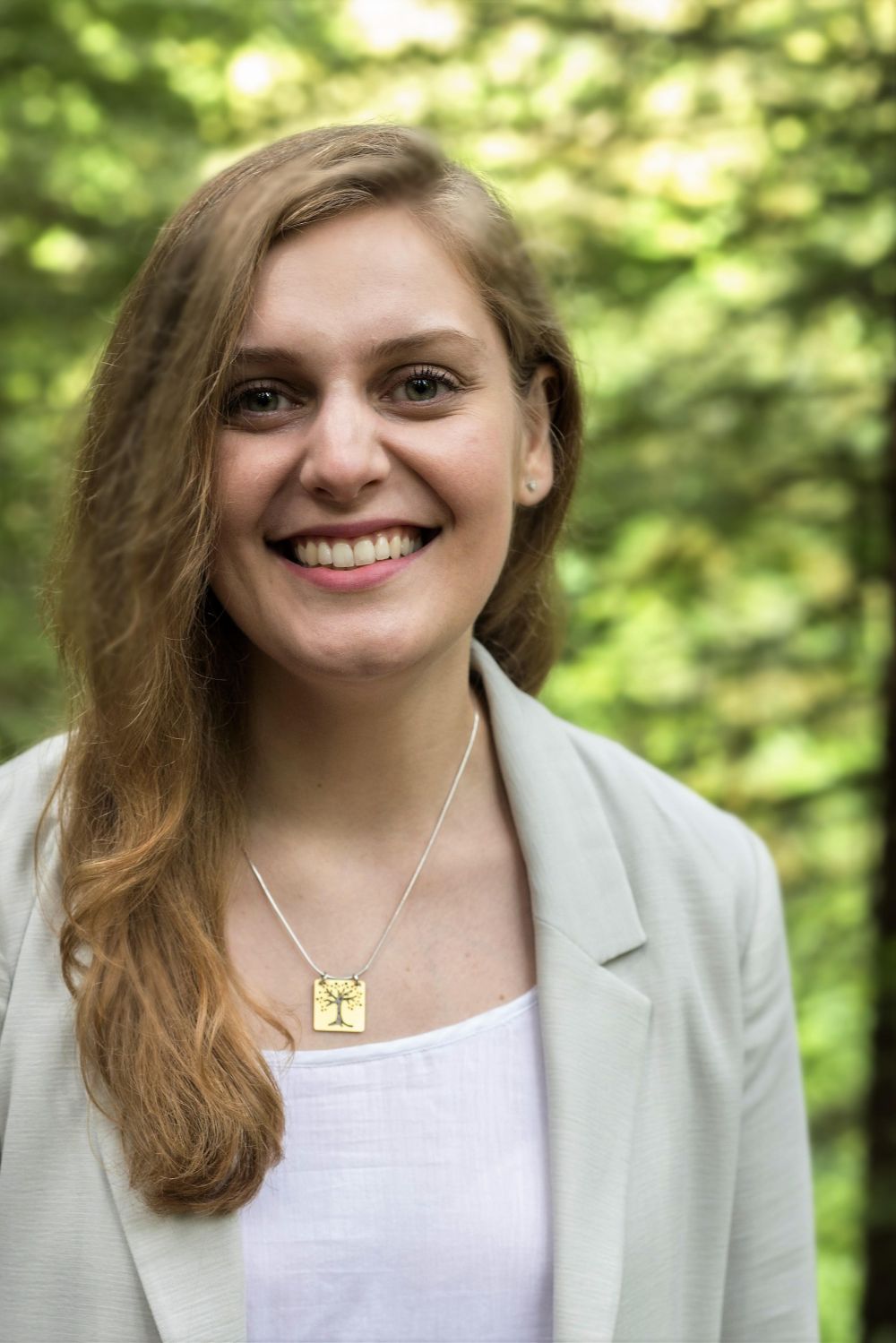 A smiling woman with (cerebral palsy and a concussion) with blazer and a tree of life necklace standing in a forest. The necklace says "whatever you can do or dream you can, begin it. Boldness has genius power and magic in it."