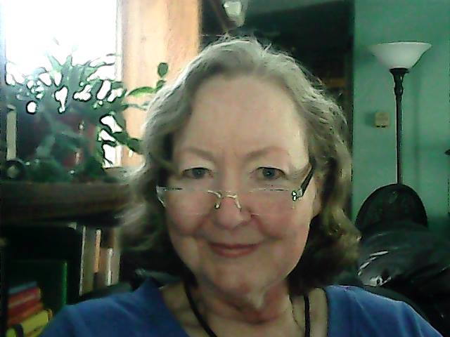 Headshot of Lynda indoors, near a window which is full of light. There's a succulent plant to her right and she looks over her glasses directly at the camera.