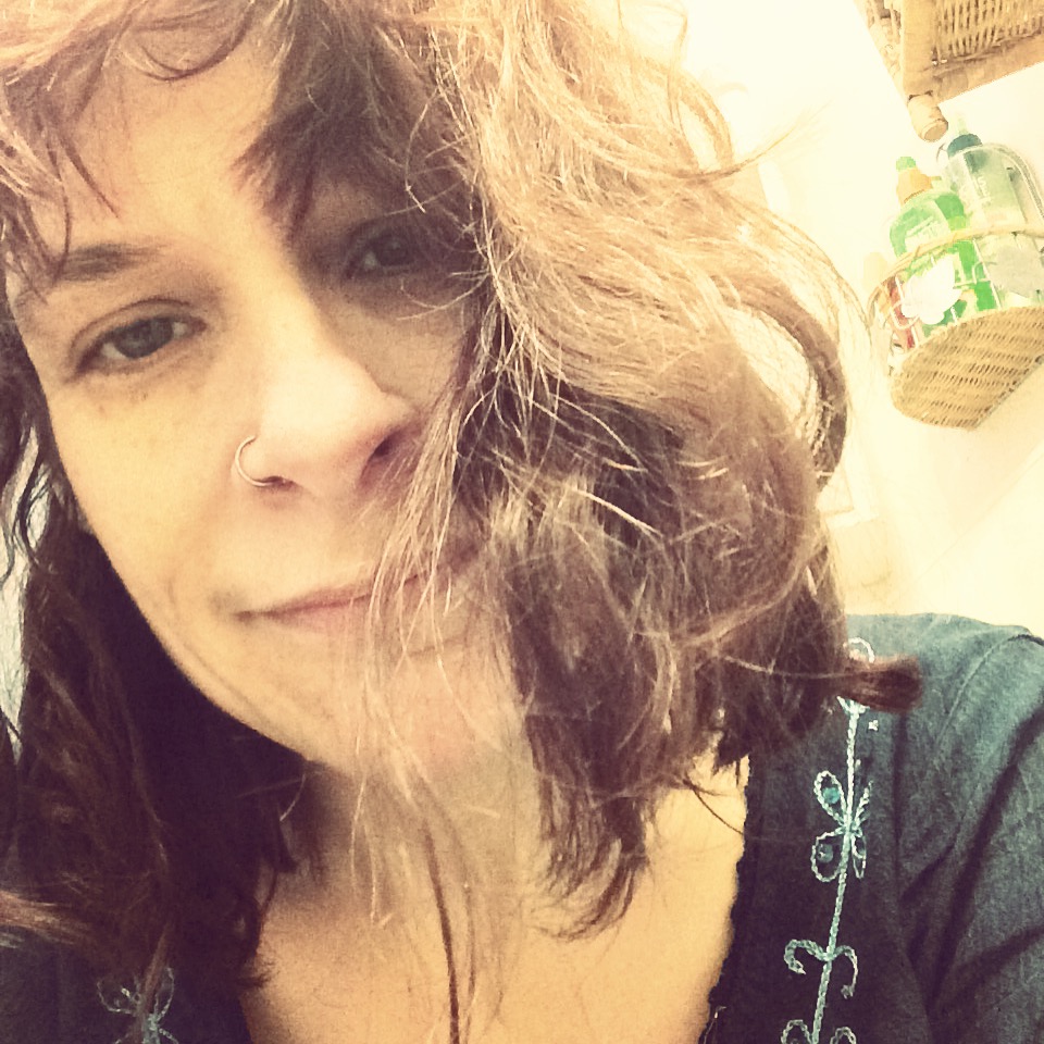 Woman in a blue top, wavy brown hair smiling sleepily into the camera.