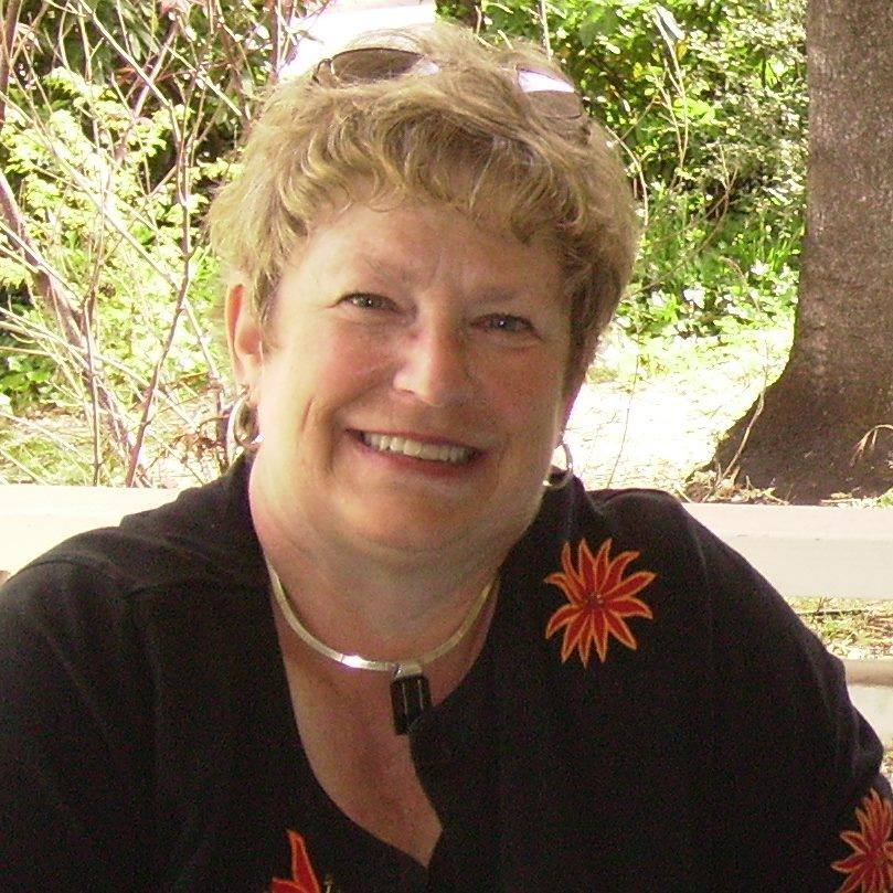Headshot of Peggy outdoors, sporting a short blonde haircut and black shirt with a red floral pattern. She's smiling and looking at the camera.