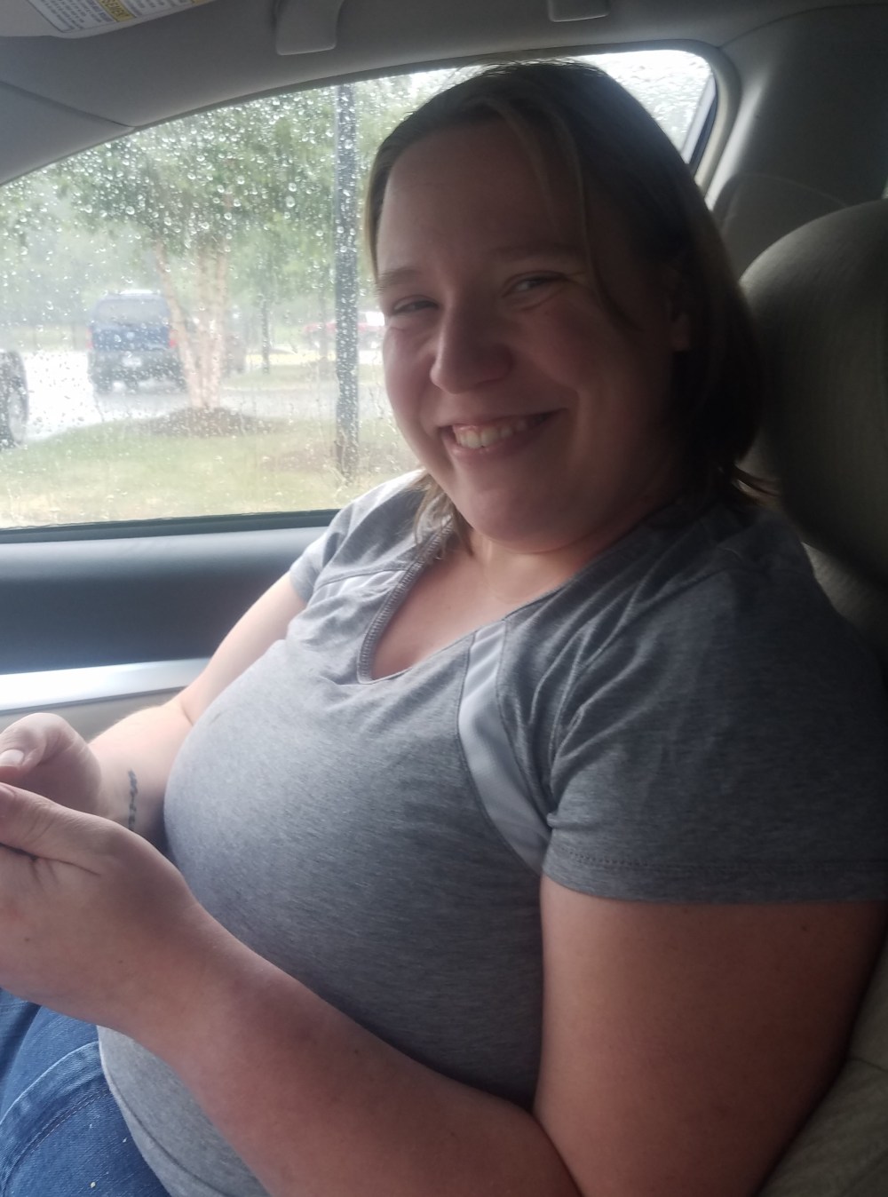 A woman with straight dark blonde hair sitting in a car with a gray shirt on. She is grinning at the photographer and the window behind her is spotted with rain.