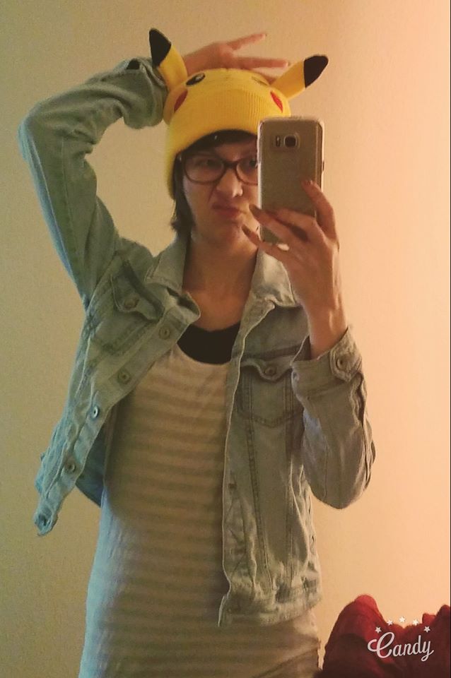 Woman holding up her phone takes a selfie in a mirror. She's wearing a jean jacket and yellow Pokemon hat.