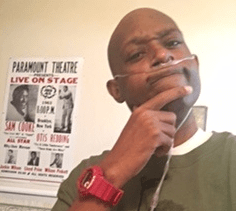 A black man with a shaved head, breathing through a cannula, wearing a green Bobba Fett shirt, a red watch, and a quizzical expression on his face. There is a vintage Paramount poster and a glimpse of a framed "Terminus" map from "The Walking Dead" in the background.