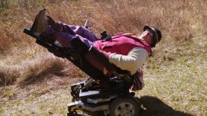 Sandra Gail Lambert—A white woman in her sixties on a nature trail. She is seated in a power wheelchair that is titled back with the feet extended into the air—a sort of hammock.