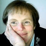 A woman with bangs smiling into the camera and resting her face in her hand. 