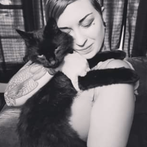 A black and white photo of a woman in a sleeveless shirt hugging a black cat, eyes closed.