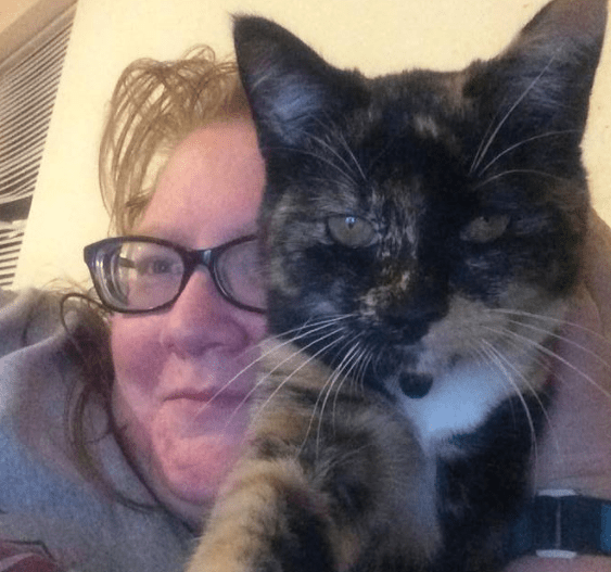 A woman with glasses wearing a gray sweatshirt, holding a tortise-colored cat in front of her.