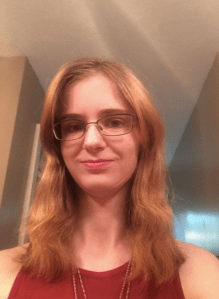 A woman with blond hair and glasses smiles and looks down into the camera, wearing a red shirt