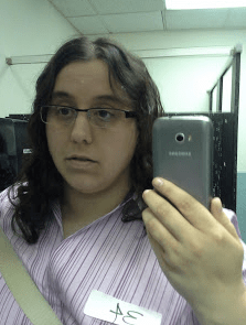 A woman with dark hair and glasses in a purple striped shirt holds up a phone to take a picture
