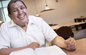 A tilted photo of a man laughing, wearing a white shirt, sitting at a table with a book open on the table, one arm on the table beside the book