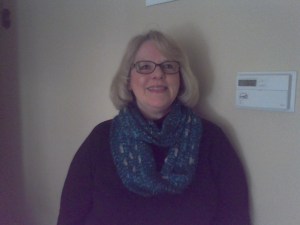 A woman in a long sleeved purple shirt and gray hair in a bob and a blue scarf around her neck. smiling