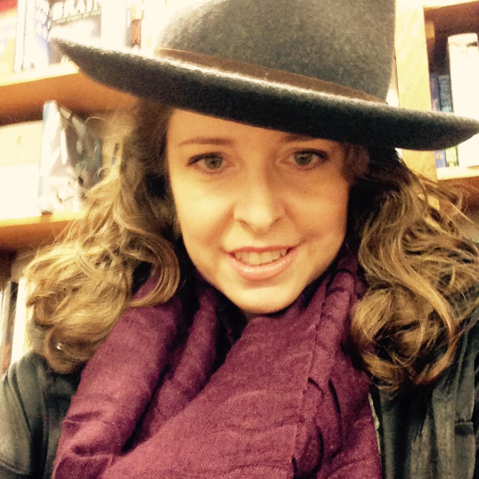 A woman with shoulder-length dark blond hair sitting in a bookstore and smiling while wearing a grey wool fedora with a brown band, a plum scarf and a black jacket. 