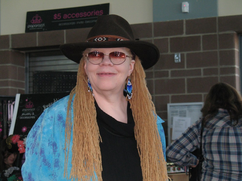 Woman with golden hair and blue earrings wears a black hat and tinted glasses.