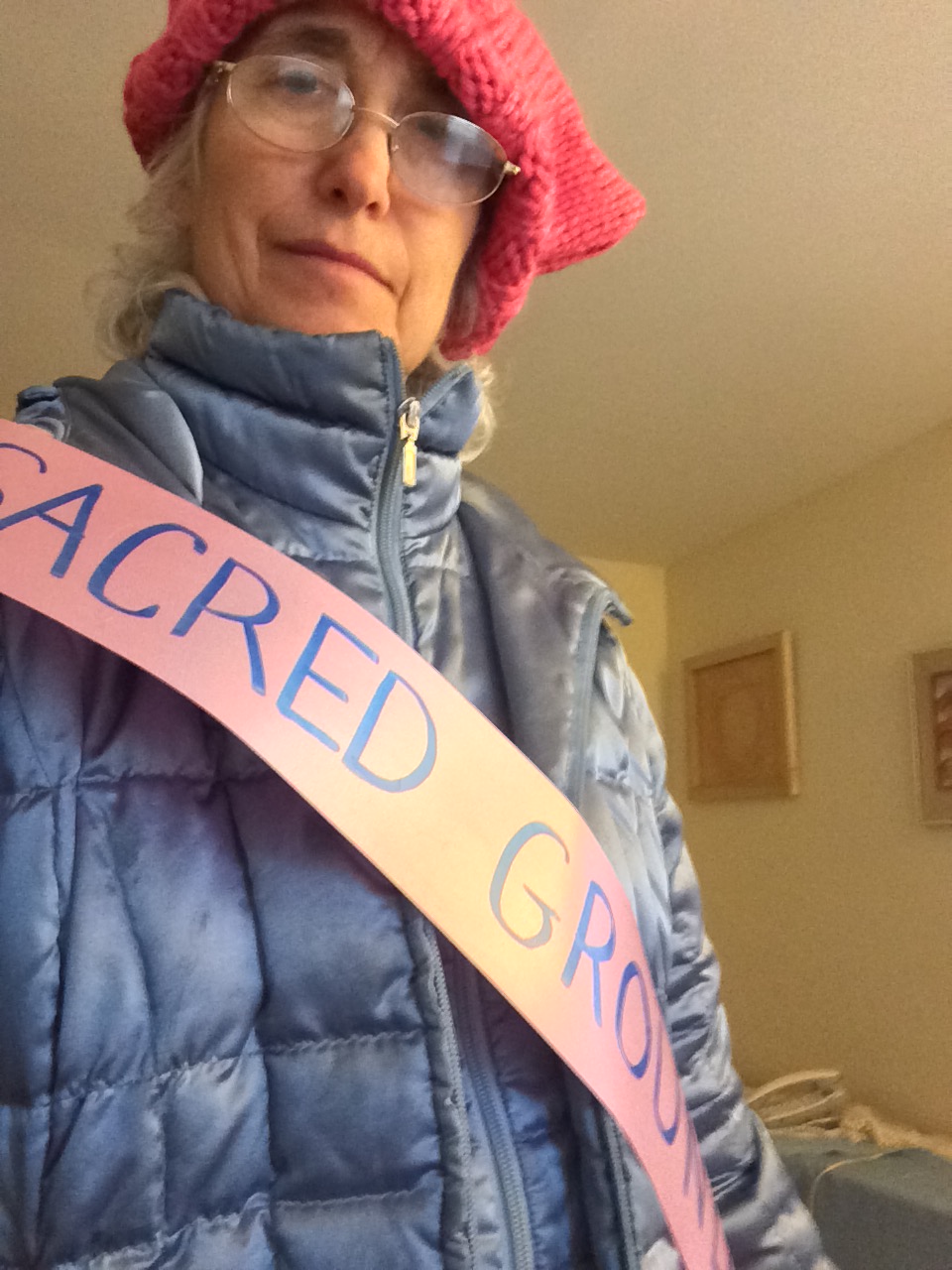 Photo taken from below, a woman looks down at the camera. She's wearing a blue winter coat, pink "pussy" hat, and a paper banner that says "SACRED GROUND."